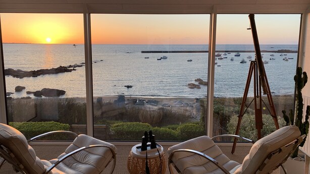 Una habitación con un ventanal grande que da a una costa rocosa y al mar al atardecer. Hay dos sillas blancas con cojines y una pequeña mesa redonda con un telescopio.