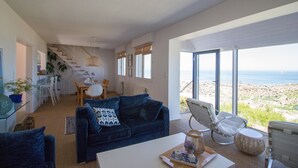Amplia sala de estar con un sofá azul, una mesa de comedor y vistas a la playa a través de amplios ventanales.