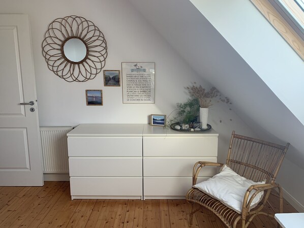 A room with a white dresser, a wicker chair with a cushion, a decorative mirror, and a framed sign with text.
