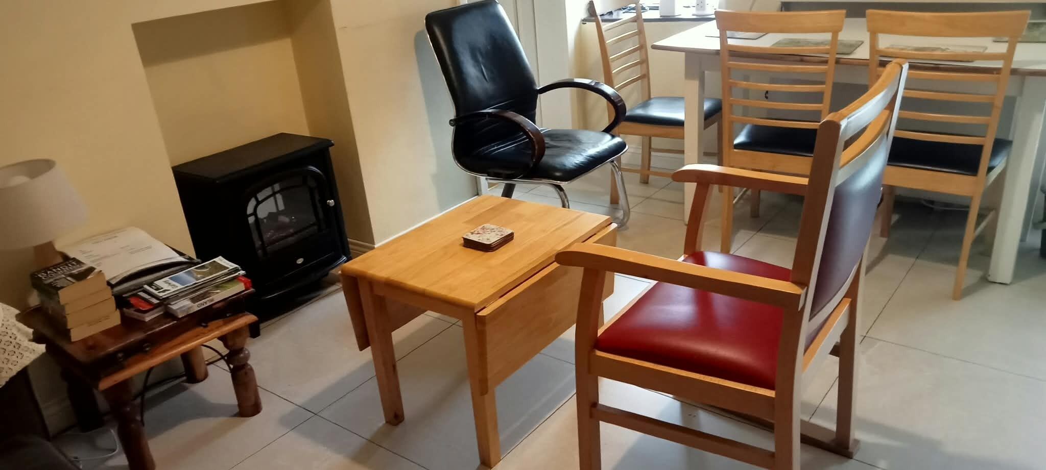 A room with a fireplace, a wooden table, a chair with red cushions, and a black leather chair.