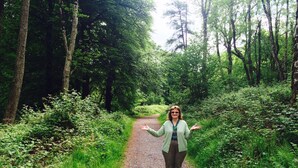 A woman standing on a gravel path surrounded by dense greenery and trees.