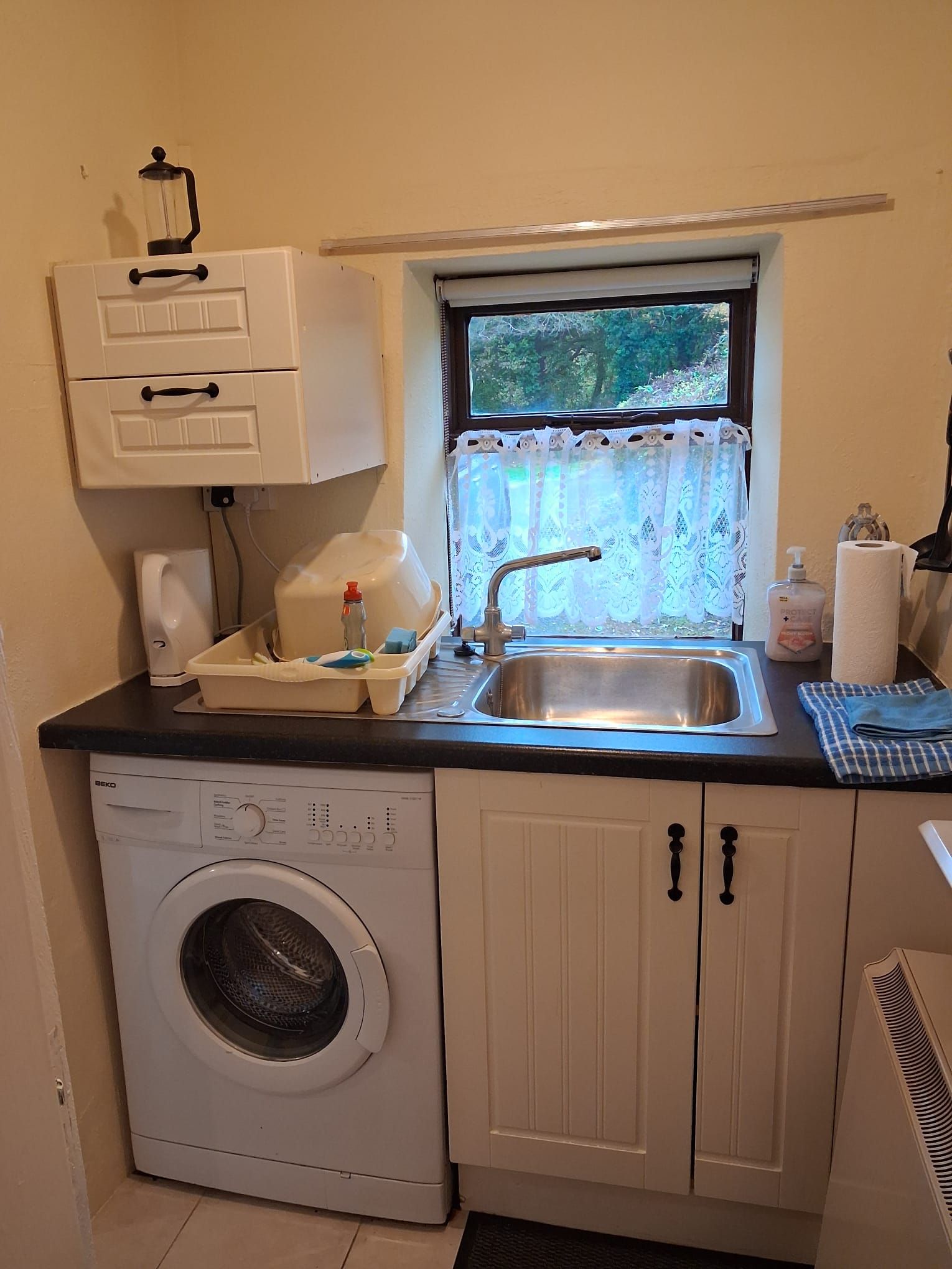 A compact kitchen with a washing machine, a sink, a window with lace curtains, and a small countertop with various items.