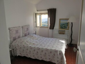 Een slaapkamer met een bed, een raam met gordijnen, een nachtkastje, een lamp en een schilderij aan de muur.