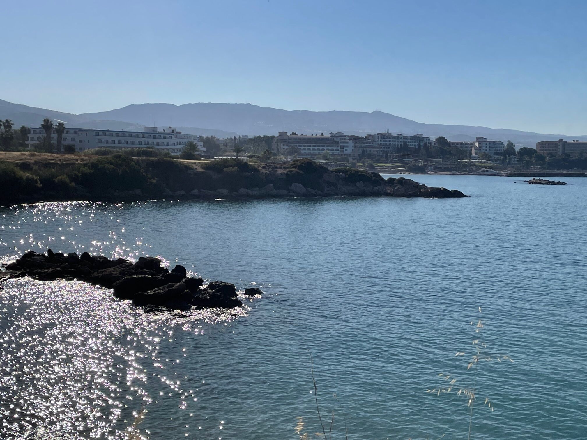 A coastal view with a rocky shoreline, calm water, and distant buildings.
