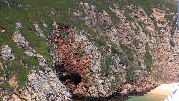 Un paysage côtier rocheux, avec un ciel bleu dégagé, une petite formation rocheuse ressemblant à une grotte, et une plage où se trouvent quelques bateaux et des espaces aménagés pour s’asseoir à l’extérieur.