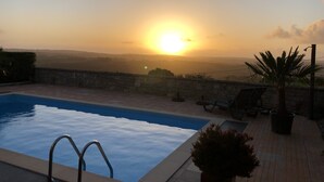 Une piscine bordée d’un mur en pierre, offrant une vue sur le coucher de soleil au-dessus d’un paysage vallonné.