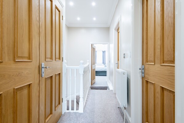 A narrow hallway with wooden doors on both sides, a white railing, and a carpeted floor leading to a bedroom with a bed and a window.