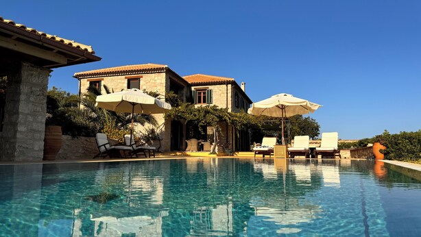 Une piscine bordée de chaises longues et d’ombrelles, un bâtiment en pierre avec un toit en tuiles et une végétation luxuriante.