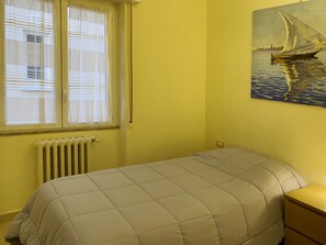 Une chambre à coucher avec un lit, une fenêtre avec des stores, un radiateur et un tableau accroché au mur.