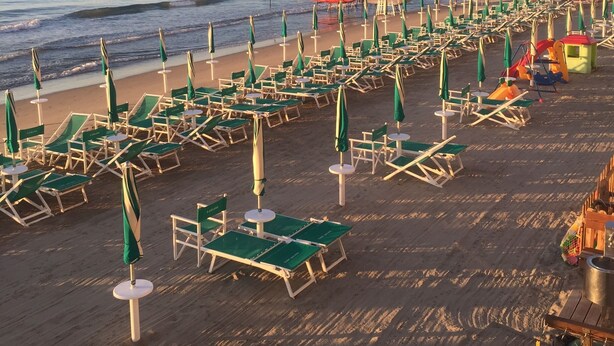 Une plage bordée de rangées de chaises longues et de parasols, une tour de surveillance pour les sauveteurs, et une vue sur la mer et les collines lointaines.