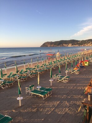 A beach with rows of sun loungers and umbrellas, a lifeguard tower, and a view of the sea and distant hills.
