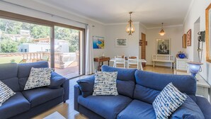 A modern living room with a blue sofa set, a dining area with a white table and chairs, and a view of the outside.