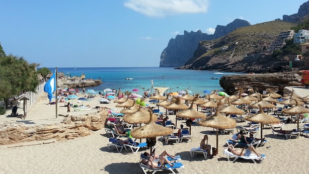 A beach with numerous sun loungers and umbrellas, a flag, and a rocky coastline.