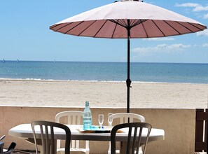 Une table entourée de chaises et protégée par un parasol, donnant sur une plage, avec l’océan et des voiliers visibles au loin.