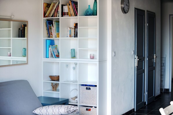 Un salon moderne avec un canapé gris, une bibliothèque blanche remplie de livres et d’objets décoratifs, une horloge murale et un couloir avec une porte.