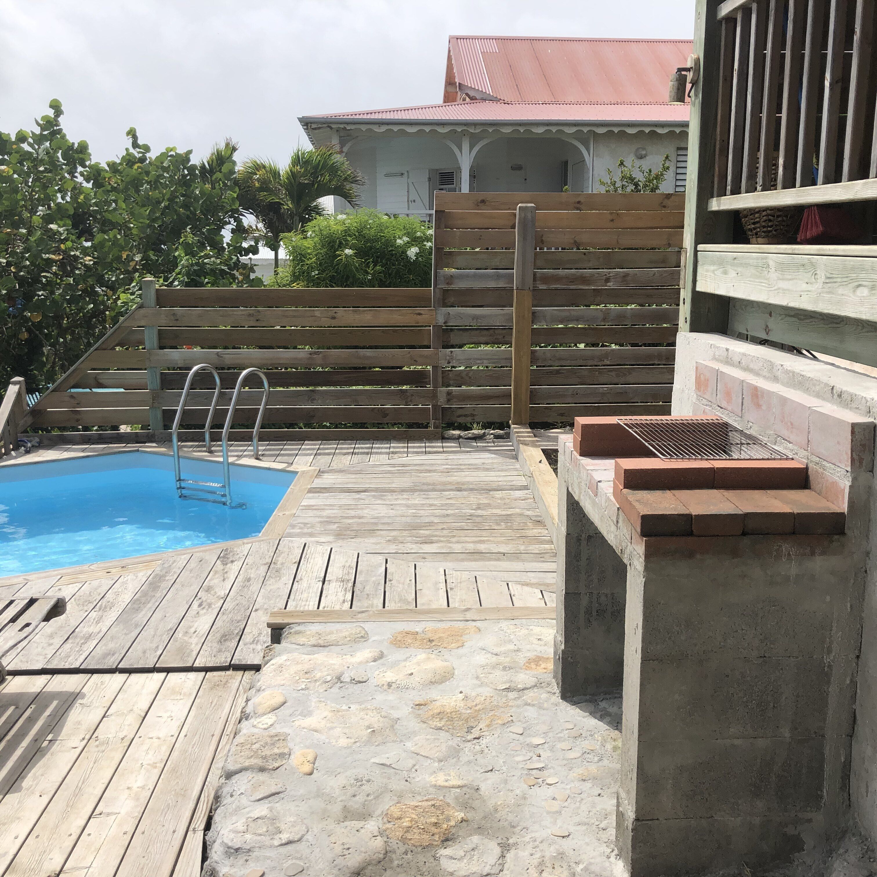 A wooden deck with a swimming pool, a brick barbecue area, and a house in the background.