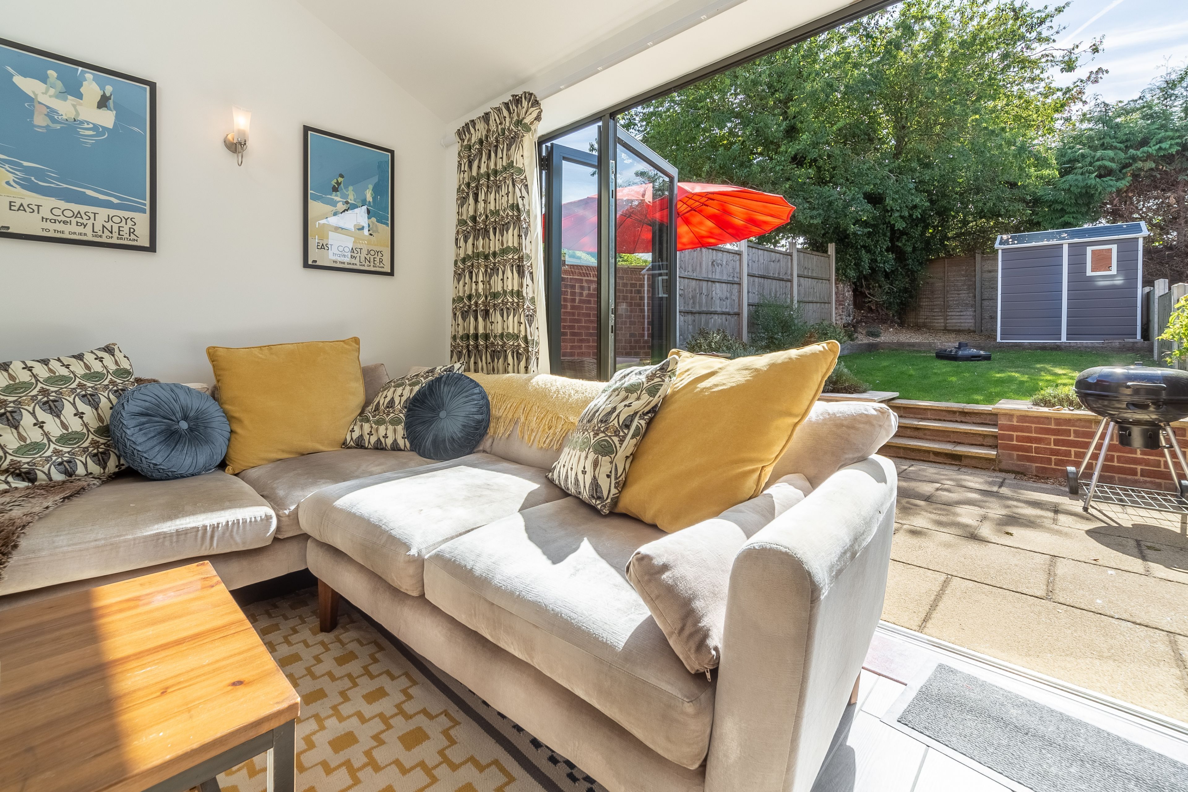 A living room with a beige sofa, patterned cushions, a wooden coffee table, and a glass door leading to a garden.
