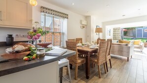 A modern kitchen with a dining area, featuring a wooden table, wicker chairs, and a large window with blinds. - Driftwood Cottage, Brancaster, Norfolk (King's Lynn)
