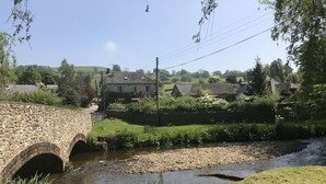 A stone bridge over a narrow stream with houses and greenery in the background. - Cosy cottage with large garden in gorgeous rural setting. (Dalwood, Devon)