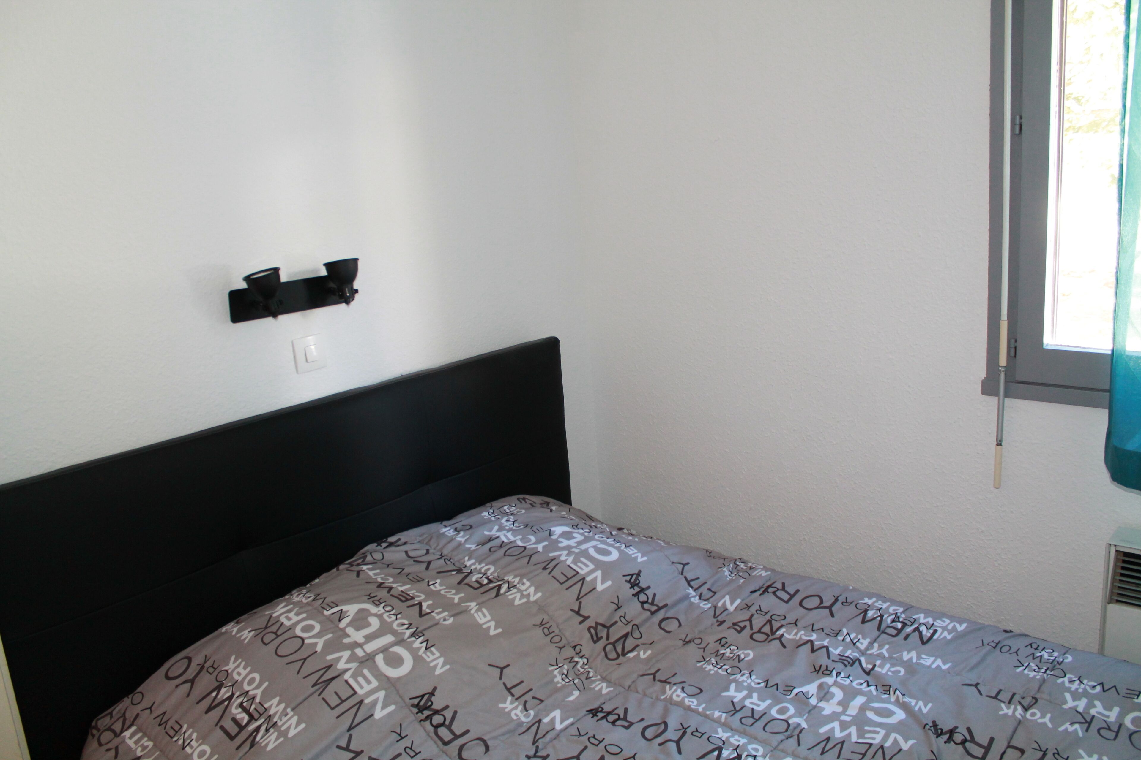 Un dormitorio con cabecera negra, cama con colcha estampada, lámpara de pared y ventana con cortina.