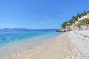 A beach with clear turquoise water, pebbly shore, and buildings on a hillside.