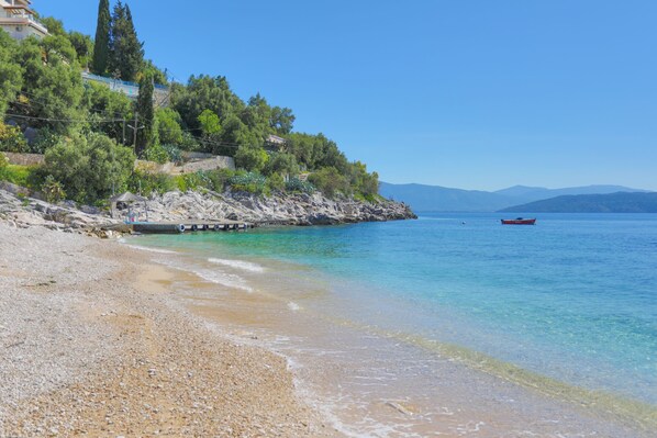 A pebble beach with clear water, a boat, and a building on a hill.