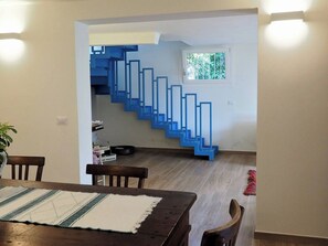 A dining area with a wooden table and chairs, a staircase with blue railings leading to an upper level, and a window with a view of greenery.