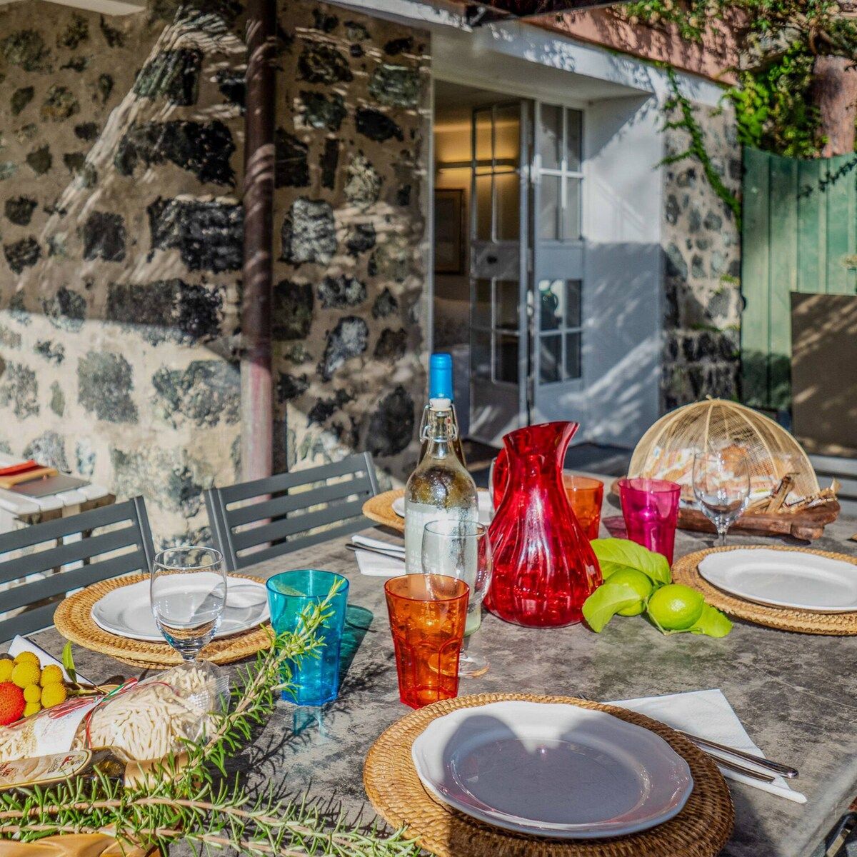 An outdoor dining table set with plates, glasses, a bottle, and a pitcher, surrounded by greenery and a stone wall.