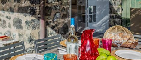 An outdoor dining table set with plates, glasses, a bottle, and a pitcher, surrounded by greenery and a stone wall.