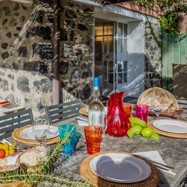 An outdoor dining table set with plates, glasses, a bottle, and a pitcher, surrounded by greenery and a stone wall.