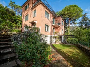 A two-story building with a stone staircase leading to a door, surrounded by lush greenery and a clear sky.