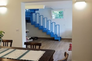 Un coin repas avec une table et des chaises en bois, un escalier avec des rampes bleues et une petite fenêtre donnant sur un paysage verdoyant.