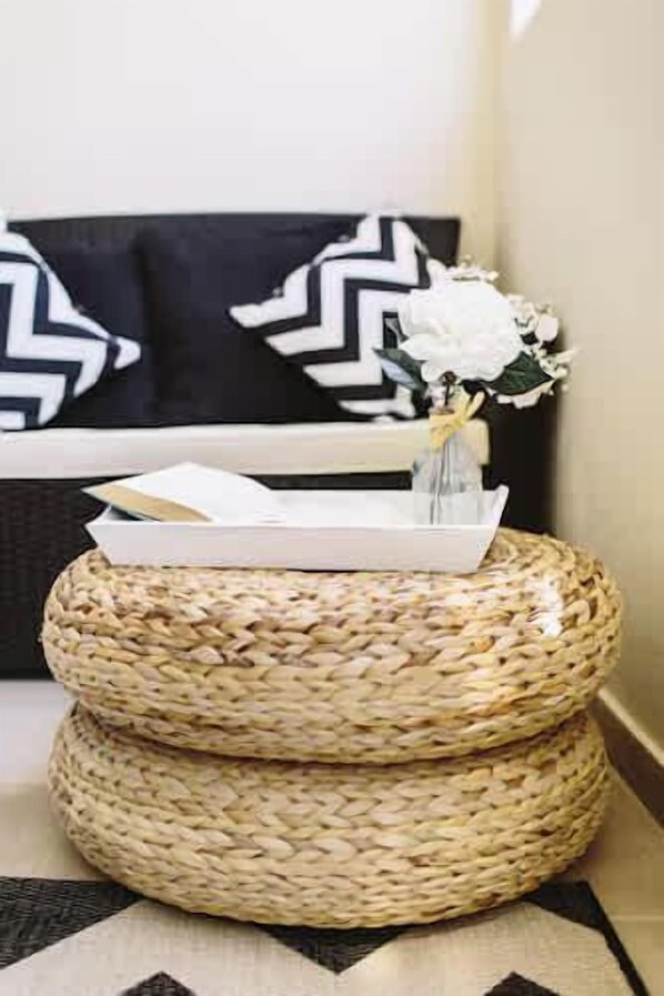 Un rincón con un taburete tejido, un jarrón con flores y un cojín blanco y negro.