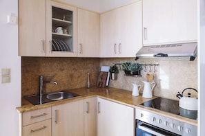 Una cocina con alacenas de madera clara, un lavamanos de acero inoxidable, una estufa y una encimera con una planta y utensilios.