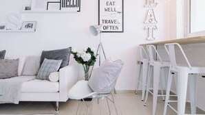 A modern living room with a white sofa, a metal chair, a dining table with white chairs, and wall art including 'Hello' and 'Eat Well Travel Often'. - Family Getaway with Pool in Southern Crete (Lagkada)