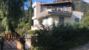 Una moderna casa a due piani con un balcone, immersa nel verde e con una montagna sullo sfondo.
