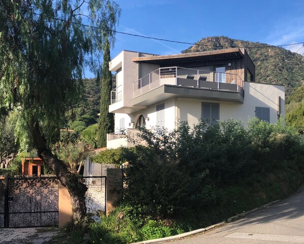 Une maison moderne à deux étages, dotée d’un balcon, entourée d’une végétation luxuriante et avec une montagne en toile de fond.