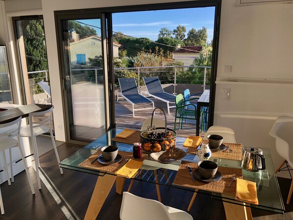 A modern dining area with a glass table, white chairs, and a view of the outdoor deck with lounge chairs.