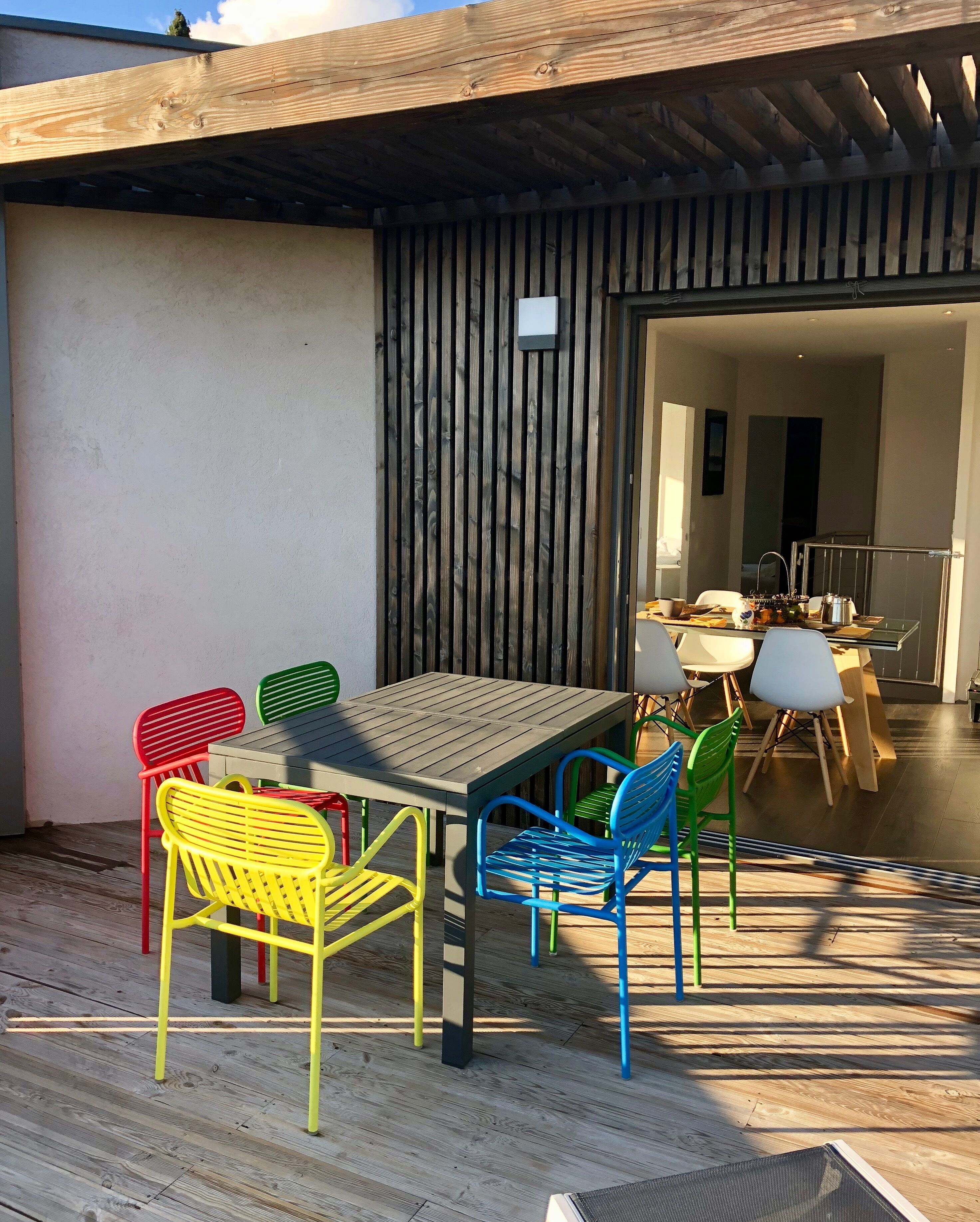 Un patio exterior con una pérgola de madera, una mesa con sillas y vista al interior.