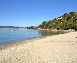 A sandy beach with a calm sea, a boat, and houses on a hillside in the background.