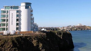Ett modernt hotell med ett cylindriskt torn och flera balkonger, beläget nära en kustlinje med utsikt över havet och avlägsna byggnader.