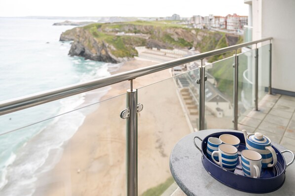 A balcony with a view of a beach and cliffs, a table with a tray of cups and a teapot, and a railing.