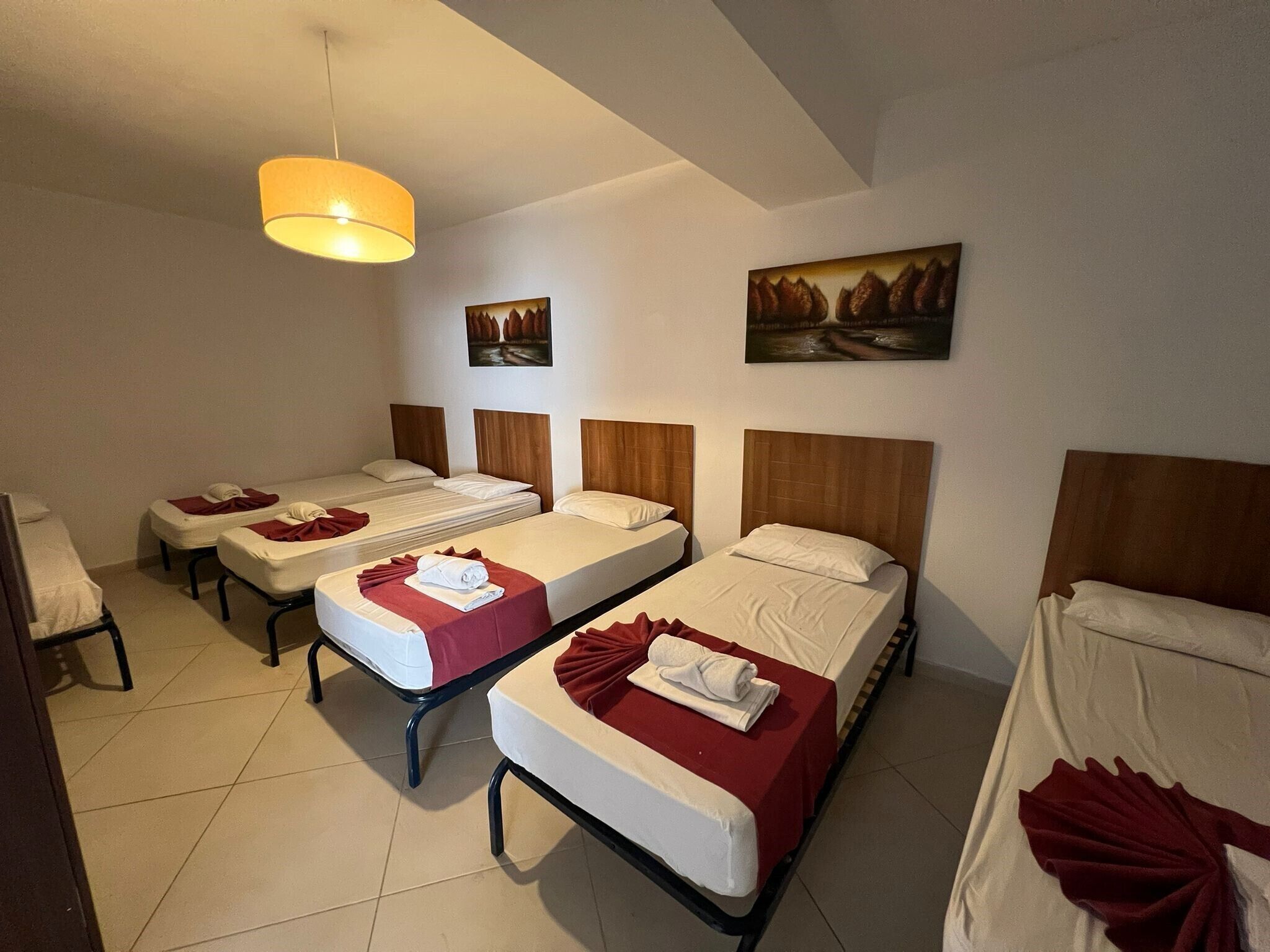Une chambre d’hôtel avec quatre lits simples, chacun recouvert d’une couverture rouge et doté d’un oreiller blanc. Il y a une grande fenêtre sur le côté gauche, un tableau accroché au mur au-dessus des lits et un luminaire suspendu au plafond.