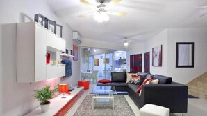 Ein modernes Wohnzimmer mit einer Couch, einem Couchtisch und einem Fernseher. Im Hintergrund ist ein Deckenventilator und eine Treppe zu sehen.