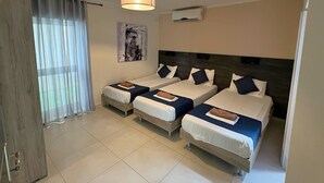 Ein Hotelzimmer mit drei Einzelbetten, einer Deckenleuchte, einer Klimaanlage und einem Fenster mit durchsichtigen Vorhängen.