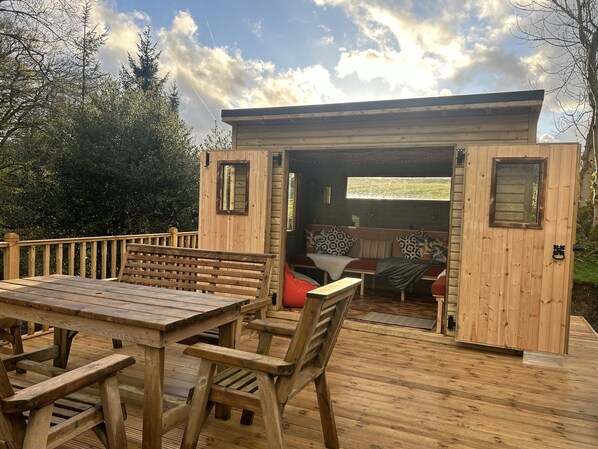 A wooden deck with a table and chairs, a bench, and a small wooden structure with a bench inside.
