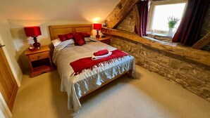 A bedroom with a stone wall, a wooden headboard, a bed with red bedding, a nightstand with a lamp, and a window with curtains. - 5* STUNNING LUXURIOUS BEAMED COTTAGE - SECLUDED PEACEFUL PEAK DISTRICT LOCATION (Leek)