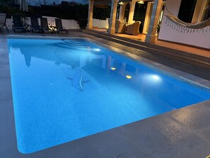 En swimmingpool med flisekant, omgivet af liggestole og et overdækket siddeområde.