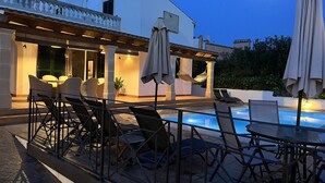 Zona de piscina a l'aire lliure amb gandules i ombrelles, una pèrgola amb cadira d'hamaca, i un edifici blanc amb un balcó.