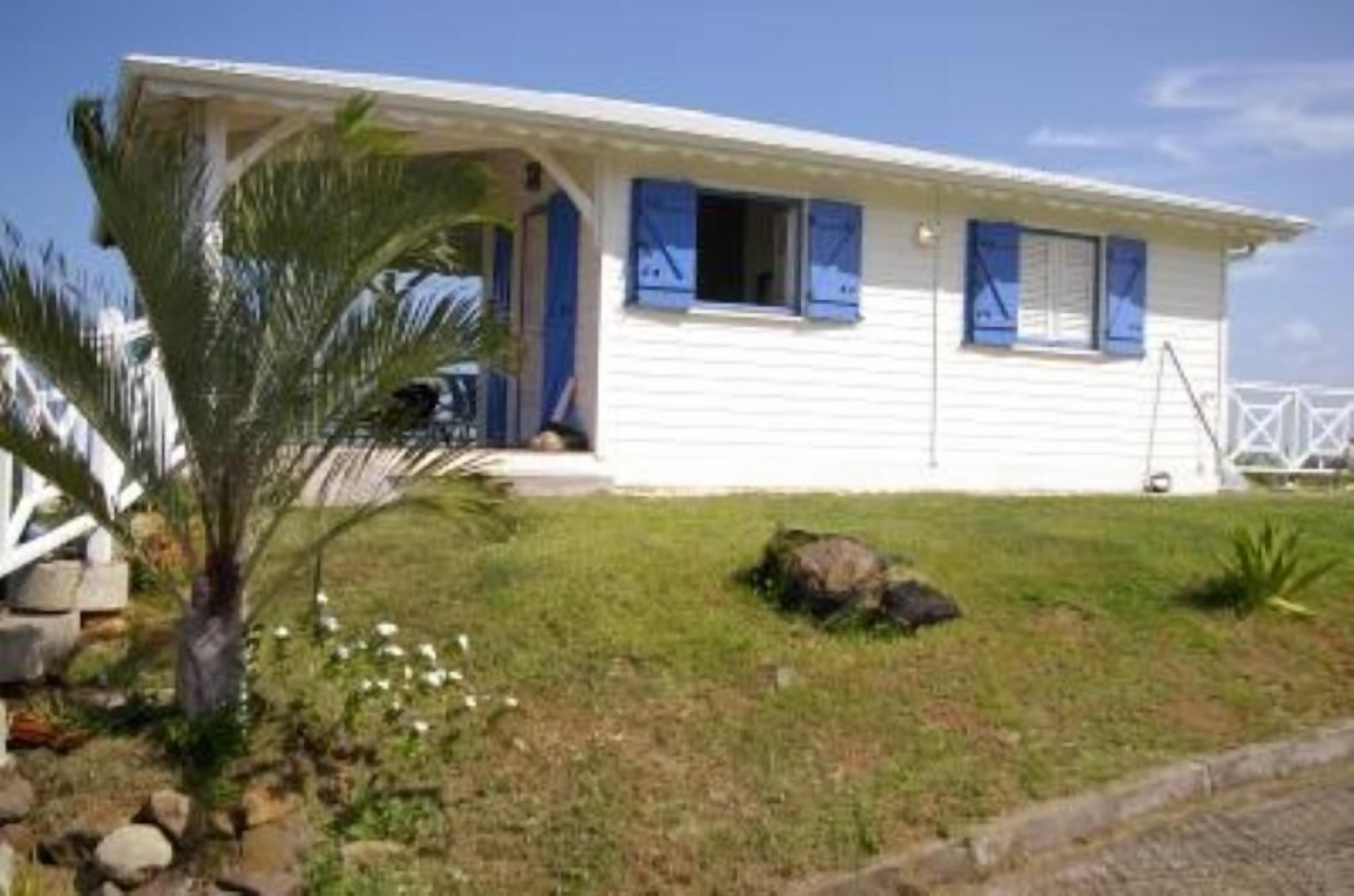 Une maison blanche d’un seul étage, avec des volets bleus, un porche et un petit jardin agrémenté d’un palmier.